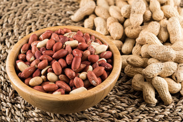 Wooden bowl of peeled peanuts and peanuts in nutshell on natural matting