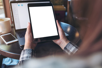 Mockup image of hands holding black tablet pc with white blank screen , laptop , smartphone and coffee cup on table in cafe