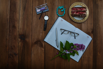 Notepad, glasses, pen, lilac branch, cosmetics, clock and cake on a wooden table. Top view, space for your text.