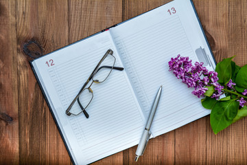 Notepad, pen, glasses and a lilac branch on a wooden table.