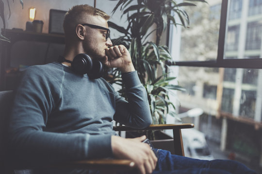 Pensive young man wearing glasses casual clothes.Man sitting in vintage armchair modern loft studio and relaxing whith headphone music.Panoramic windows on blurred background.Horizontal.