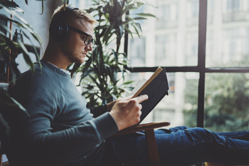 Portrait handsome young man wearing glasses casual clothes holding book hands.Man sitting in vintage armchair modern loft studio reading book. Blurred background.Horizontal.