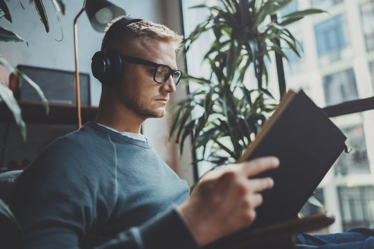 Handsome young man wearing glasses casual clothes.Man sitting in vintage armchair modern loft studio, reading book and relaxing whit headphone music. Blurred background.Horizontal.Cropped.