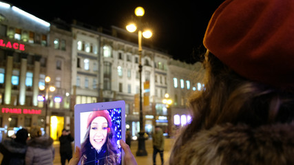 Attractive young girl taking pictures of herself on the screen in the background of the Christmas tree in the evening in the city