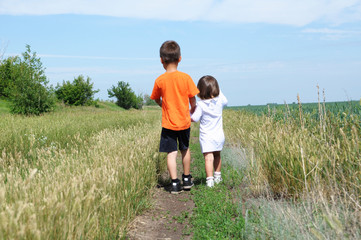 Little boy and little girl walking away on the road in the field at summer day, brother and sister...