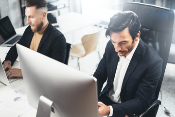 Two elegant mans working at sunny office on desktop computer while sitting at wooden table.Blurred background,horizontal.