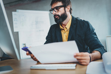 Young bearded man working at sunny while sitting at wooden table.Designer analyze new ideas on modern computer.Blurred background,horizontal.