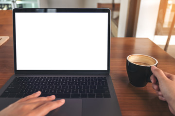 Mockup image of a woman using laptop with blank white screen while drinking coffee on wooden table in office