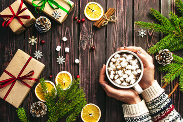 Female hands in woolen sweater with a cup of hot coffee with marshmallows, Christmas gifts, spruce branches, cones, orange slices, snowflakes. Traditional Christmas dark background top view. 