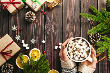 Female hands in woolen sweater with a cup of hot coffee with marshmallows, Christmas gifts, spruce branches, cones, orange slices, snowflakes. Traditional Christmas dark background top view. 
