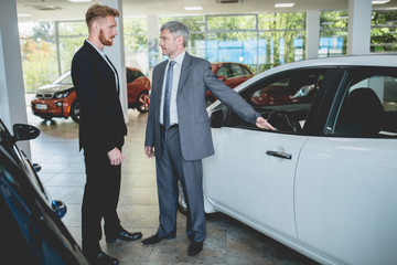 Young man at auto showroom talking with vehicle dealer. Man choosing electro car in car salon.