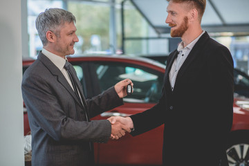 Car seller and buyer shaking hands in auto showroom. Young bearded man buying new vehicle.