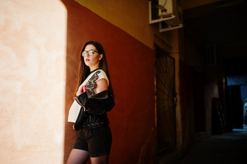 Fototapeta premium Stylish brunette girl wear on leather jacket and shorts with glasses against orange wall at shadows.