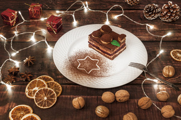 Wooden table with lights, white plate with Christmas cake, walnuts, dried orange, anise and red gifts