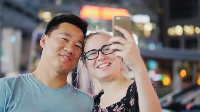 Young multi-ethnic couple in the night city among the lights of advertising. Tourists photograph the attractions of the concept