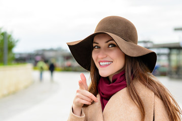 Attractive woman in a brown hat and beige coat looking at the camera points the finger at you and smiles. Pick me.