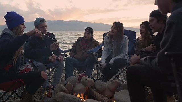 Group of millenials gathered around campfire at sunset next to lake roasting marshmallows.