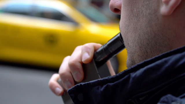 Young hipster millennial man smoking vape e-cigarette outside on New York City street during day time. Wearing blue jacket, yellow taxi cab passes by in Manhattan past office building break time