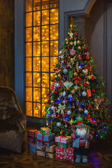 Decorated with toys and Christmas lights, the Christmas tree stands in the room. Under the Christmas tree gifts. Next to the tree is a sofa with a rug. The window is covered with New Year's garlands