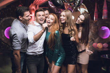 Young people in the club dance and sing. A man in a white shirt is holding a microphone and singing.