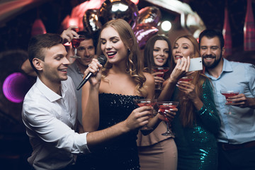 A woman in a black dress is singing songs with her friends at a karaoke club. Her friends do selfie.