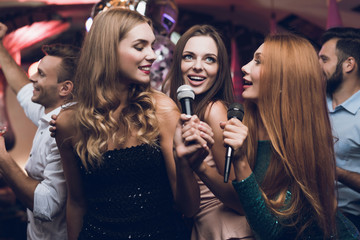 Three beautiful girls sing in a karaoke club. Behind them are men waiting for their turn.