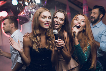 Three beautiful girls sing in a karaoke club. Behind them are men waiting for their turn. - obrazy, fototapety, plakaty