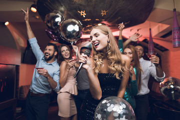 Young people have fun in a nightclub and sing in karaoke. In the foreground there is a woman in a black dress. - obrazy, fototapety, plakaty