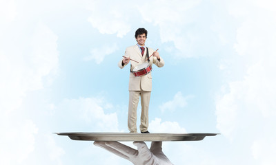 Young businessman on metal tray playing drums against blue sky background
