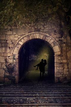 Silhouette of a couple, man holding a woman in his arms, walking through dark arch with yellow backlight. Stone castle wall of an Old Town.
