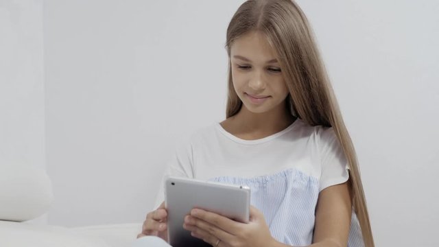 Cute girl sitting on sofa with tablet computer doing homework and playing games