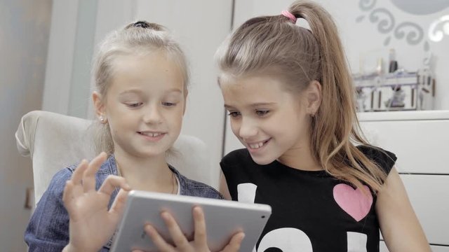 Two beautiful little sisters sit at a table and play on a Tablet PC. Happy smiling girls