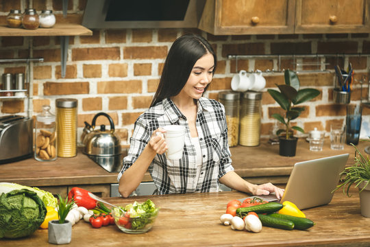 Young Beautiful Girl With Laptop On Kitchen Finding Recipes And Smiling. Food Blogger Concept