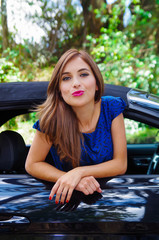 Close up of beautiful woman wearing a blue dress and posing inside of a luxury black car on a roadtrip. the car standing on the sidelines, in a blurred nature background