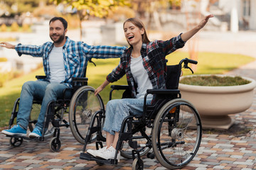 A man and a woman on wheelchairs ride around the park. They put their hands to one side and fooled around.