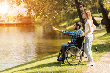 Fototapeta na wymiar A woman is walking in the park with a man in a wheelchair. They are on the shore of a lake.