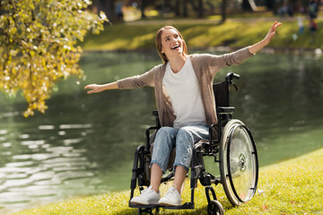 A woman in a wheelchair sits on the shore of a lake. She spread her arms out and laughed.