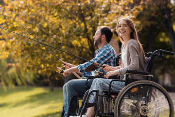A woman and a man in wheelchairs are sitting on the lake shore. They have spinners in their hands...