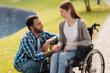 A woman is sitting in a wheelchair. A man is sitting next to her and holding her hands.