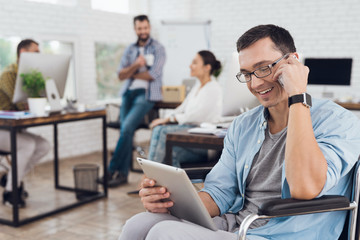 Disabled person in the wheelchair works in the office. He talks on smartphone.