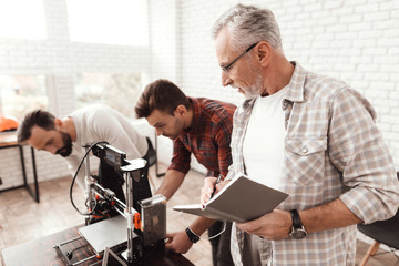 Three men set up a self-made 3d printer to print the workpiece. An elderly man with a notebook...