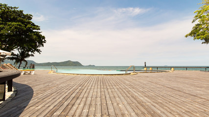 Fototapeta premium Swimming pool having Natural wooden floor platform which located beside the clean and clear water in ocean, with white cloud and blue skyas background at skyline.