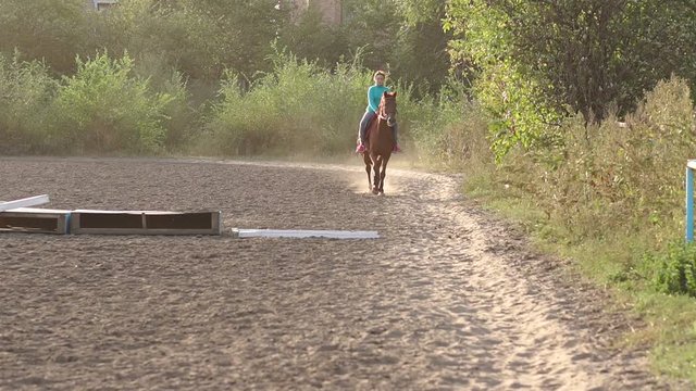 Teenage girl with excess weight rides a brown horse on a horse farm at sunset. Slow motion.