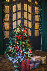 Decorated with toys and Christmas lights, the Christmas tree stands in the room. Under the Christmas tree gifts. Next to the tree is a sofa with a rug. The window is covered with New Year's garlands