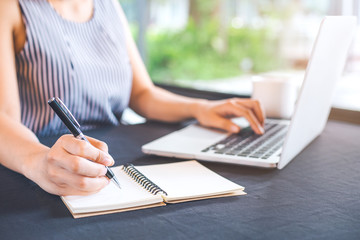 Woman hand writing on notepad with a pen and works in a laptop computer in the office.