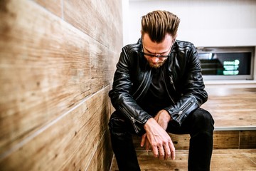 Handsome man with a beard in a black jacket and sunglasses sitting on the stairs with his head down. Men's beauty, fashion. Business-like style.