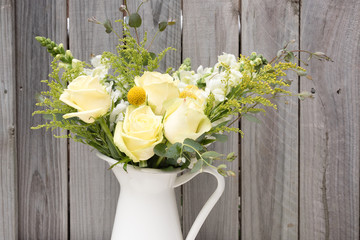 Close up of beautiful bouquet of spring flowers including yellow roses and craspedia with green foliage in white jug against rustic grey fence (selective focus)