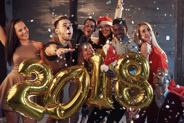 Obraz na płótnie Canvas New 2018 Year is coming! Group of cheerful young multiethnic people in Santa hats carrying gold colored numbers and throwing confetti on the party