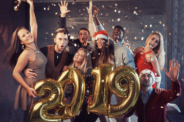 New 2019 Year is coming! Group of cheerful young multiethnic people in Santa hats carrying gold colored numbers and throwing confetti on the party