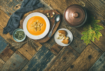 Fall warming pumpkin cream soup with croutons and seeds on board over wooden background, copy space, flat-lay, top view. Autumn vegetarian, vegan, healthy comfort food cooking and eating concept